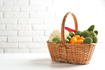 Different fresh raw vegetables in wicker basket on white wooden table against brick wall, space for text