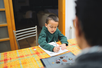 Boy concentrating while drawing or writing in a notebook at a table, receiving guidance or assistance from his blurred grandmother, highlighting intergenerational learning and family education