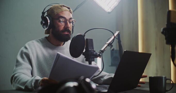 Professional Male Podcast Host Recording Episode in Studio. Bearded Man Wearing Glasses and Headphones Reads From Script Into Microphone in Audio Studio, Focusing on Production Digital Media Content.