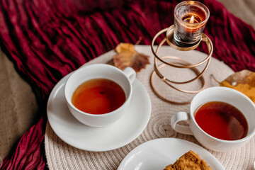 White porcelain cups with black tea on a burgundy tablecloth, candles glowing softly behind a slice of cake for a relaxed cozy mood.