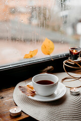 Cozy rainy-day scene with black tea on wooden sill, candle burning softly near the wet window.
