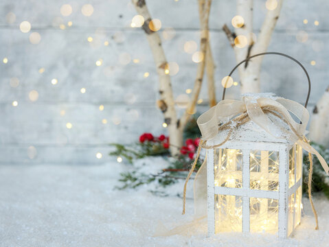 Background for personalized Christmas card. White metal lantern on snow with bokeh, birch tree branches and festive holiday twinkle fairy lights, greenery and red berries on white wood plank backdrop.