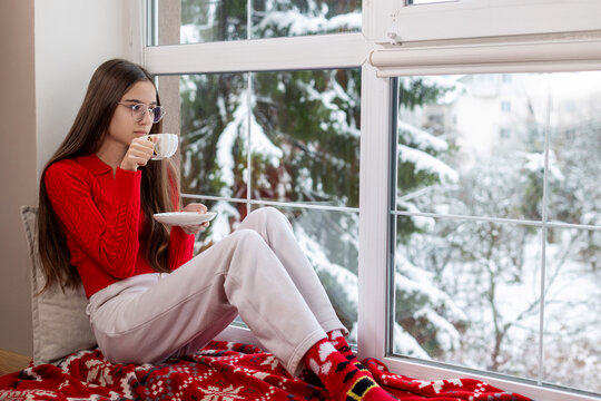 Teen girl relaxing with tea by snowy window. A young woman in red sweater and Christmas socks sipping tea on a cozy windowsill with a snowy winter view outside.