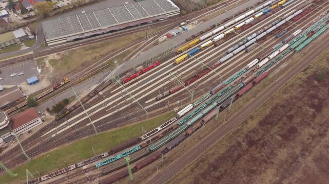 An angled aerial view of a large freight railyard featuring numerous parallel tracks filled with cargo trains, tankers, and container wagons. One train leaving the station.