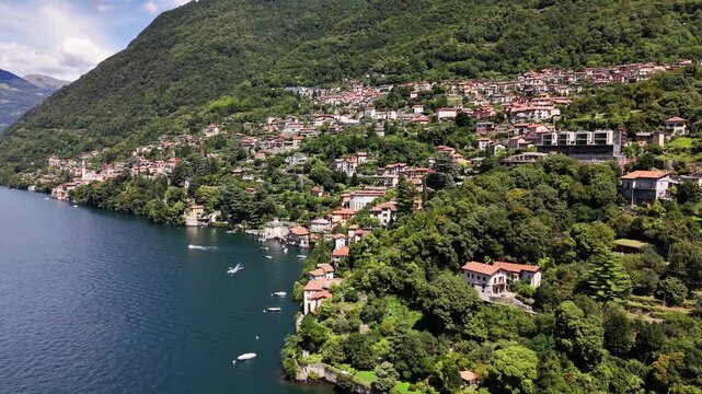 Nesso village on Lake Como's shoreline shows charming traditional Italian architecture climbing the lush green mountainside, with boats navigating the calm blue water, Lombardy, Italy