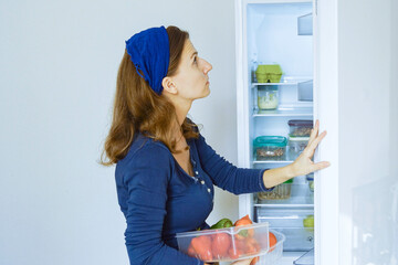 Person with a vegetable container in her hands looking up at refrigerator shelves assessing stored food items. Represents thoughtful menu planning moment and refrigerator content evaluation.