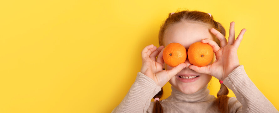 Smiling girl holding two oranges over her eyes against a bright yellow background, creating a fun and cheerful composition.