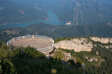 Obraz premium Circular viewing platform (mirador) with white railing, built high on a cliff, offering a stunning panoramic view of a reservoir and forested mountains near Berga, Catalonia