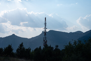 Backlit silhouette of a tall telecommunications tower with antennas and dishes, set against a mountainous landscape with sunbeams piercing through the clouds.