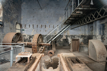 Main engine room of an abandoned cement factory featuring large, rusted hydraulic turbine components, machinery trenches, and an industrial metal staircase.