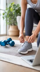 Person tying running shoes beside a laptop on a mat, with dumbbells in the background, embodying a modern home fitness lifestyle
