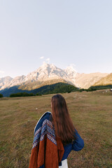 Fototapeta premium Woman with long hair from back wearing patterned shawl standing in meadow facing mountain landscape. Nature scene with wide grass field, outdoor travel and peaceful horizon.