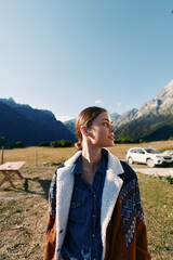 Fototapeta premium Woman portrait in mountains near parked car on meadow, wearing warm jacket and denim shirt, travel and nature scene with clear sky, outdoor lifestyle and peaceful scenery