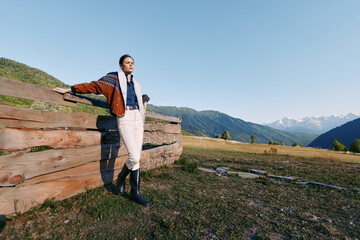 Fototapeta premium Woman stands by wooden fence in mountain meadow at sunrise, wearing jacket, scarf and rubber boots. Landscape view with rolling hills and clear blue sky for outdoors leisure.