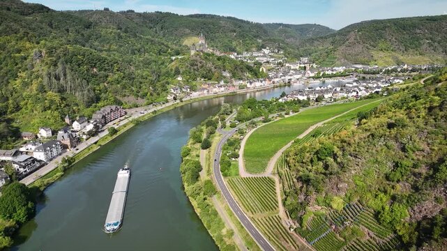 Drone shot pulling in on bend of the Moselle River, framed by lush vineyards, riverside town of Cochem and hilltop Reichsburg Castle. Bulk cargo barge sails along the Moselle town Cochem.
