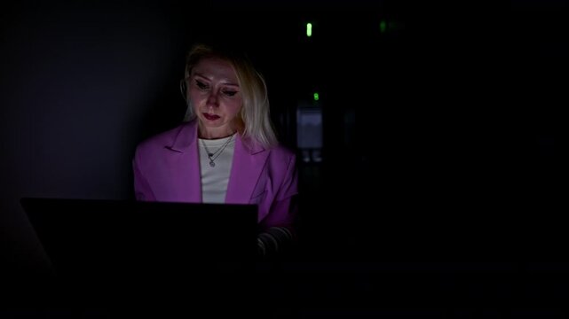 Businesswoman works diligently on her laptop in a dark server room. The screen's glow highlights her concentration, symbolizing late-night work, IT challenges, and dedicated technology professionals.