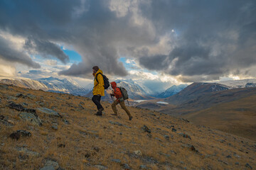 A guy and girl in bright clothing with backpacks hike across a mountain slope from right to left during autumn golden hour. A beautiful mountain valley unfolds behind them.