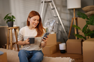 A woman relaxes with a coffee and smartphone, smiling as she navigates her new home filled with moving boxes and greenery, capturing a moment of transition and comfort.