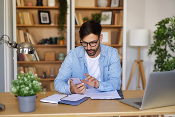 A man in a light blue shirt uses a smartphone while seated at a wooden desk in a well-organized home office, with a laptop and notebook nearby in a bright, inviting setting.