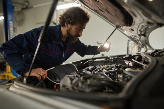 Mechanic checks engine components thoroughly with flashlight in a well-lit automotive repair shop. Tools scattered nearby, focused on repair process.