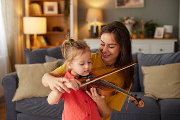 A mother smiles while guiding her young daughter in playing the violin. The family is having fun together in their comfortable living room, filled with warmth and love.