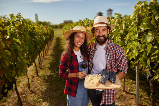 A couple smiles as they hold a tray of cheese and grapes while standing among lush vines. The sun shines bright, creating a perfect day for wine tasting in their vineyard.