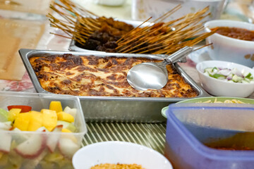 A table filled with assorted dishes including a cheesy baked lasagna in a metal tray, satay, fruit salad, sliced vegetables, sauces, and soups. 