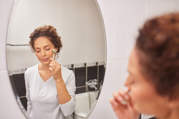 In a bright bathroom, a woman gently applies a skincare product to her face while looking at her reflection in the mirror. It is part of her daily morning routine focused on self-care.