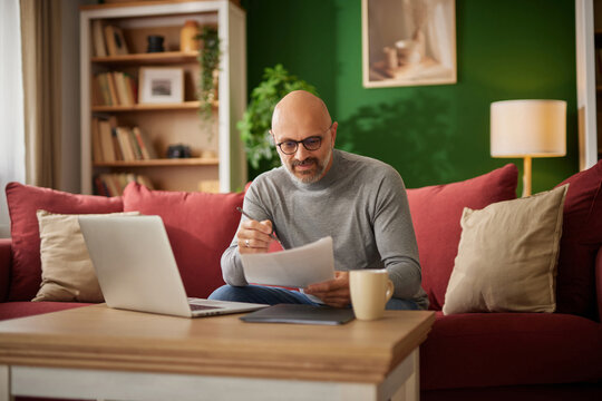 A man sitting on a red couch reviews financial documents while using a laptop in a well-lit living room. A warm atmosphere enhances his concentration. - Powered by Adobe