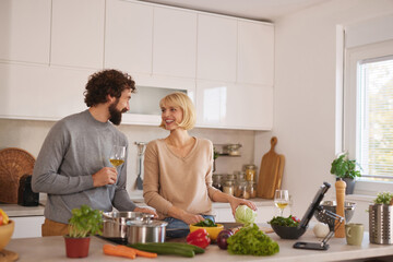 A couple prepares a meal in a modern kitchen, surrounded by various fresh vegetables and cooking tools. They share smiles and a glass of wine, enjoying their time together.
