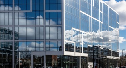 A modern glass office building with reflective windows, featuring a blue sky with white clouds.