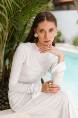 Woman in a long white dress sitting and posing gracefully beside tropical greenery and a pool