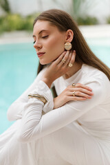 Woman in a white dress posing by a pool with her hand near her face, wearing bold earrings and rings
