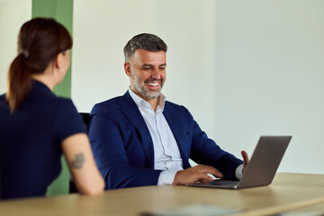 Smiling Business Professional Using Laptop At Office With Female Colleague In Discussion