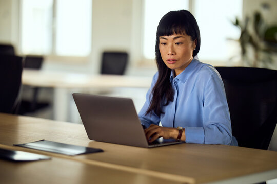 Asian Woman Working on Laptop in Modern Open Office Space, Focused Professional at Desk Today