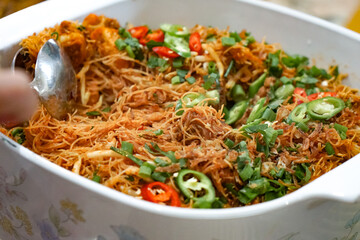 A large white dish of stir-fried rice vermicelli with vegetables, crispy shallots, and sliced chili peppers, served at a buffet setting.