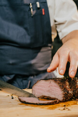 A chef is cutting a piece of meat on a wooden cutting board. The knife is positioned above the meat, and the chef's hand is holding the knife