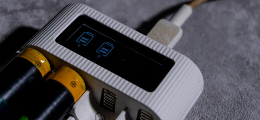 Close-up of a battery charger displaying two charging indicators on its digital screen, with AA batteries inserted. Dramatic lighting highlights the device?s texture and power