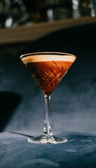 A martini glass filled with a brown liquid sits on a blue surface. The glass is almost full, with only a small amount of liquid left at the top. The drink appears to be a martini
