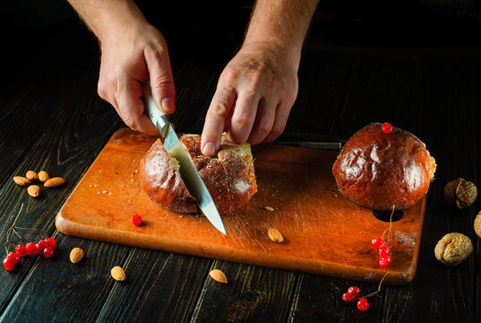 Freshly baked bread is being sliced on a wooden board by hands holding a sharp knife. Nuts and bright red berries are scattered around creating a warm kitchen atmosphere