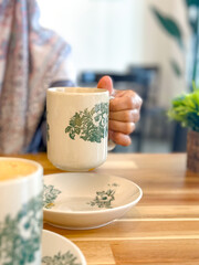 Close-up of a hand holding a ceramic mug with green floral design, surrounded by matching saucers and a potted plant on a wooden table.
