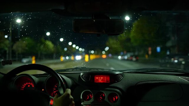 Car interior view of driving through a city streets at night. Unrecognizable male driver, blurry wide angle view of the street and tail lights, rain drops on the windshield, real time video