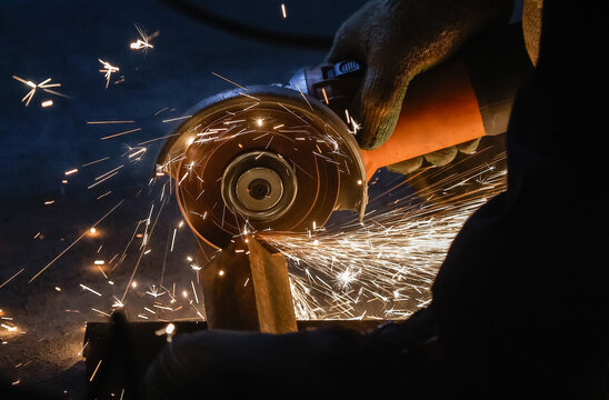 A worker uses a grinder to cut metal in a workshop. Sparks fly as the tool makes contact, illuminating the dark surroundings. The focus is on the hands and the grinding activity.