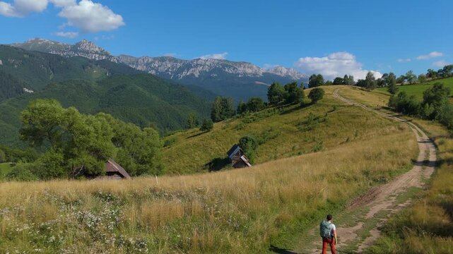 Static aerial shot of lone hiker walking away along a dirt cart road on Carpathian hills near Simon, with Bucegi Mountains ridge in the summer background