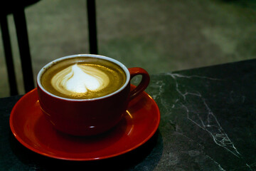 close-up of a cup of coffee with a heart-shaped latte art design. The coffee is served in a red ceramic cup placed on a matching red saucer, which sits on a dark marble table. Selective focus