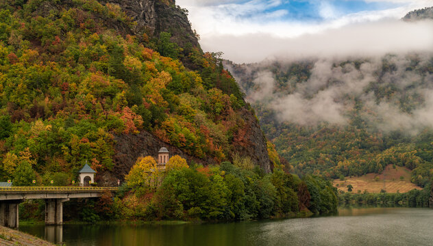 Picturesque autumn landscape in the Olt Valley, Romania, with vibrant fall foliage on the mountains. A small historic church and a bridge span the calm Olt River, surrounded by misty, forested hills.