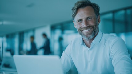 Fototapeta premium Confident Businessman with Beard Smiles at Desk in Modern Office Setting with Laptop and Colleagues in Background