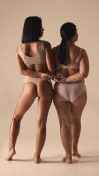 Rear view of two female models in soft neutral underwear holding hands in gentle connection. Concept of inclusive fashion storytelling, intimate branding, body-positive visuals, diversity promotion.