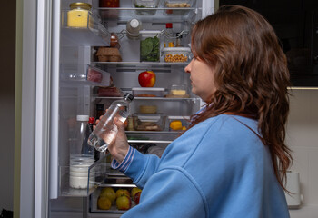 A young woman stands at an open refrigerator filled with healthy foods, containers, fruits, and