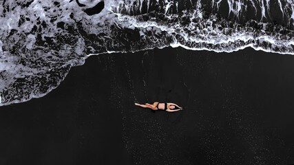 Woman relaxing on black sand beach, waves gently lapping around her, aerial view captures serene moment, camera slowly zooms out revealing expansive ocean